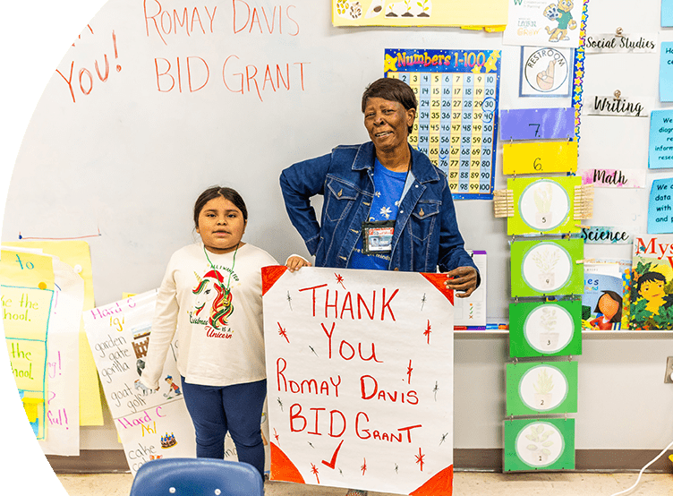 A young girl and an older woman standing in front of a whiteboard that reads "Thank You! Romay Davis BID Grant." Both are holding signs that also express gratitude for the Romay Davis BID Grant. The setting appears to be a classroom, with colorful posters and educational materials in the background.