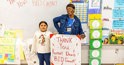 A young girl and an older woman standing in front of a whiteboard that reads "Thank You! Romay Davis BID Grant." Both are holding signs that also express gratitude for the Romay Davis BID Grant. The setting appears to be a classroom, with colorful posters and educational materials in the background.