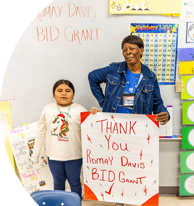 A young girl and an older woman standing in front of a whiteboard that reads "Thank You! Romay Davis BID Grant." Both are holding signs that also express gratitude for the Romay Davis BID Grant. The setting appears to be a classroom, with colorful posters and educational materials in the background.