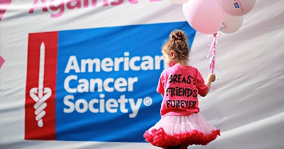 Young girl holding pink balloons. She is wearing a pink tutu and a pink shirt that says "Breast Friends Forever". Behind her there is the American Cancer Society® logo placed on a banner.