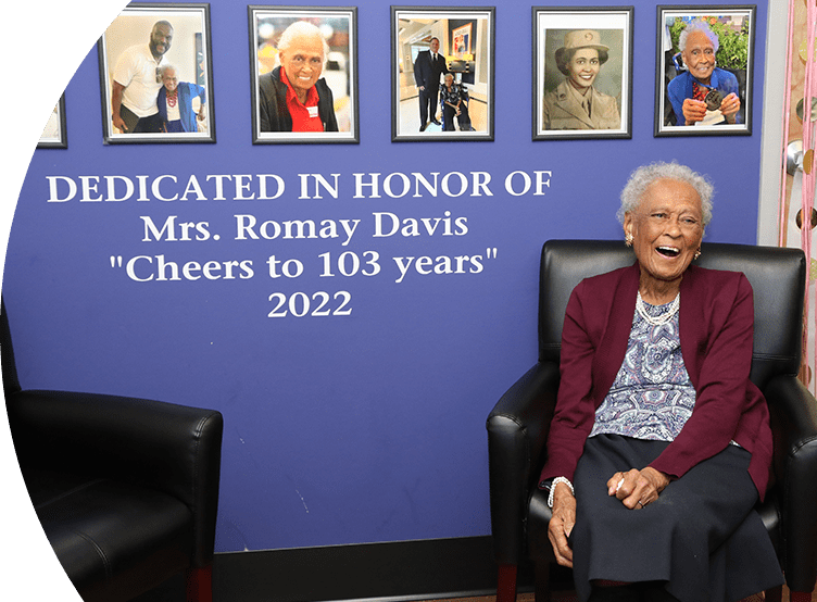 Mrs. Romay Davis joyfully sitting on a chair smiling. Behind her is a wall with framed images of her and the words "DEDICATED IN HONOR OF Mrs. Romay Davis 'Cheers to 103 years' 2022".