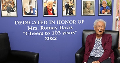 Mrs. Romay Davis joyfully sitting on a chair smiling. Behind her is a wall with framed images of her and the words "DEDICATED IN HONOR OF Mrs. Romay Davis 'Cheers to 103 years' 2022".