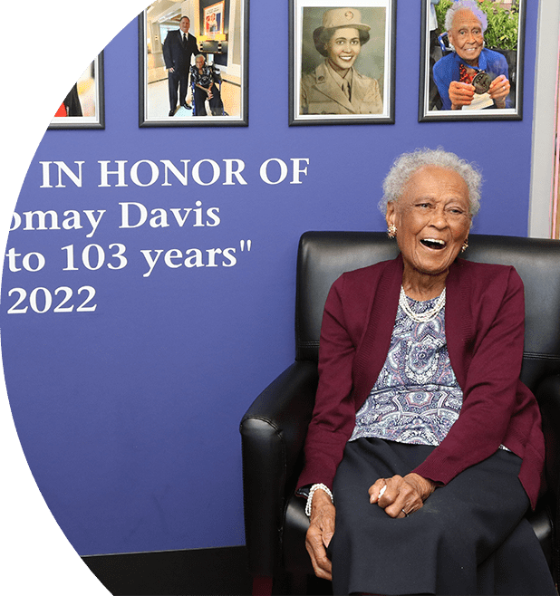 Mrs. Romay Davis joyfully sitting on a chair smiling. Behind her is a wall with framed images of her and the words "DEDICATED IN HONOR OF Mrs. Romay Davis 'Cheers to 103 years' 2022".