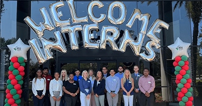 Group of interns standing in front of the Southeastern Grocers building. Above them are letter-shaped balloons with the words "Welcome Interns".