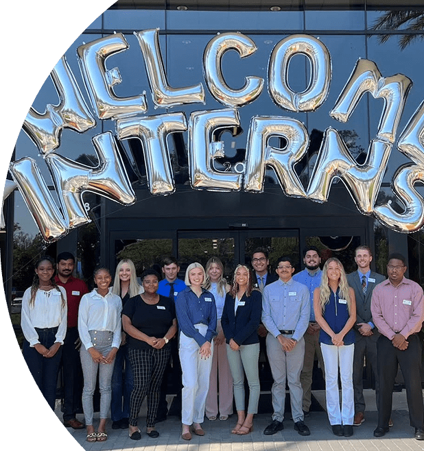 Group of interns standing in front of the Southeastern Grocers building. Above them are letter-shaped balloons with the words "Welcome Interns".