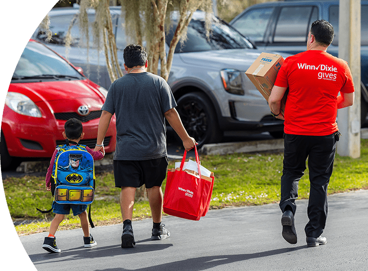 Dad holding son's hand while holding Winn-Dixie Gives Foundation bags. Next to them is a Winn-Dixie Gives Foundation volunteer helping them carry items.