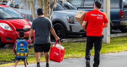 Dad holding son's hand while holding Winn-Dixie Gives Foundation bags. Next to them is a Winn-Dixie Gives Foundation volunteer helping them carry items.