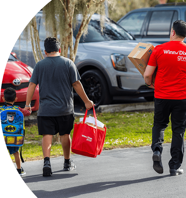 Dad holding son's hand while holding Winn-Dixie Gives Foundation bags. Next to them is a Winn-Dixie Gives Foundation volunteer helping them carry items.