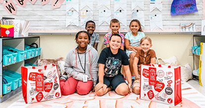 Group of school kids smiling. They are in a classroom sitting on a mat. Next to them are two Winn-Dixie Community Bags.