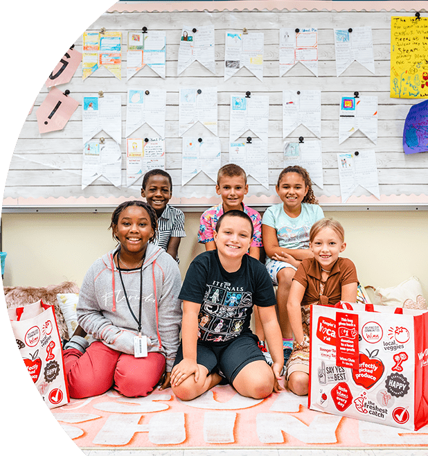 Group of school kids smiling. They are in a classroom sitting on a mat. Next to them are two Winn-Dixie Community Bags.