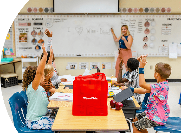 Teacher in the front of class pointing at a whiteboard. In front of her, young students are sitting on desks and raising their hands. On one of the desks there is a red Winn-Dixie reusable bag.