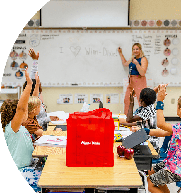 Teacher in the front of class pointing at a whiteboard. In front of her, young students are sitting on desks and raising their hands. On one of the desks there is a red Winn-Dixie reusable bag.