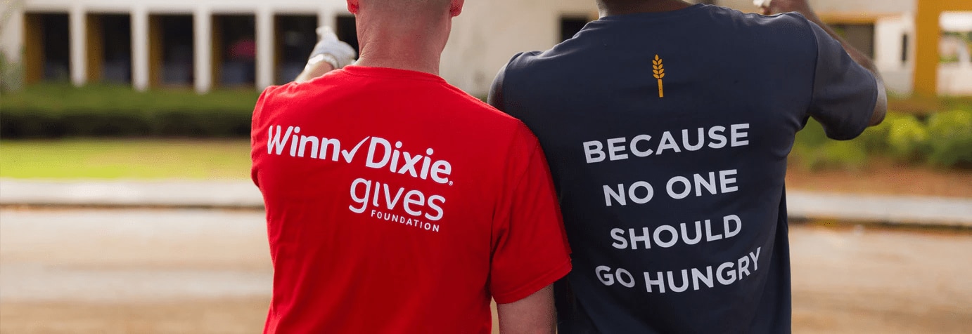 Volunteer wearing Winn-Dixie Foundation on the left next to volunteer on the right wearing a shirt with the text that reads "Because no one should go hungry".