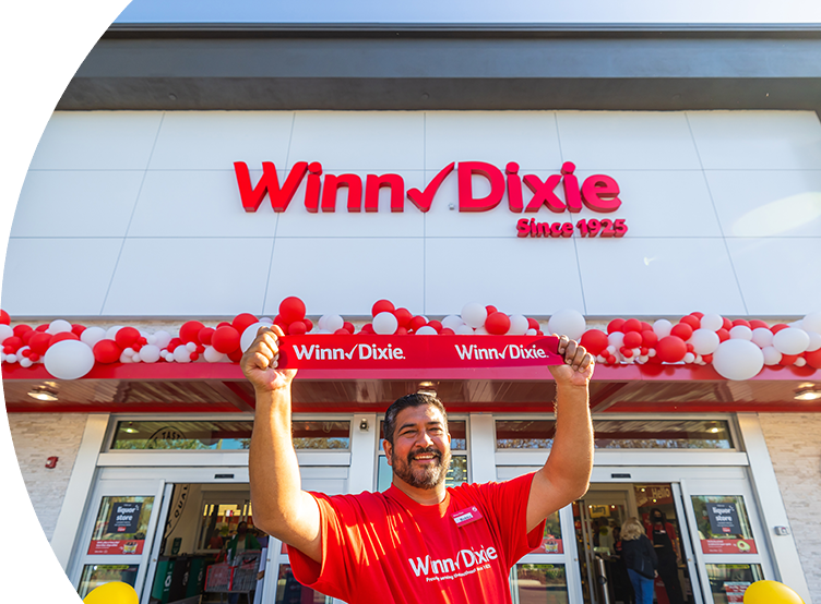 Joyful employee holding up a Winn-Dixie sign in front of a Winn-Dixie store