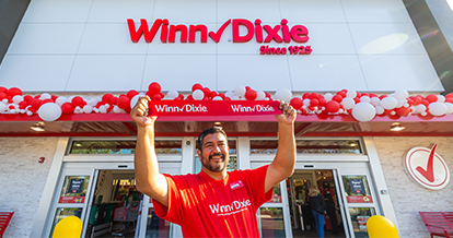 Joyful employee holding up a Winn-Dixie sign in front of a Winn-Dixie store