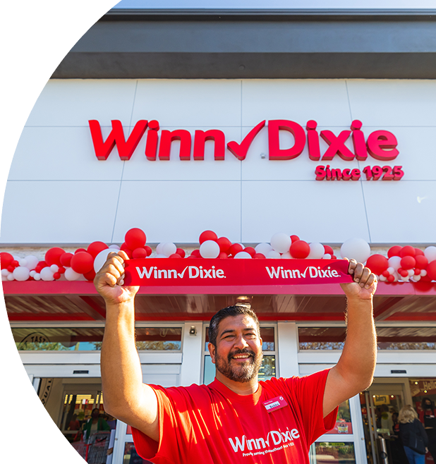 Joyful employee holding up a Winn-Dixie sign in front of a Winn-Dixie store