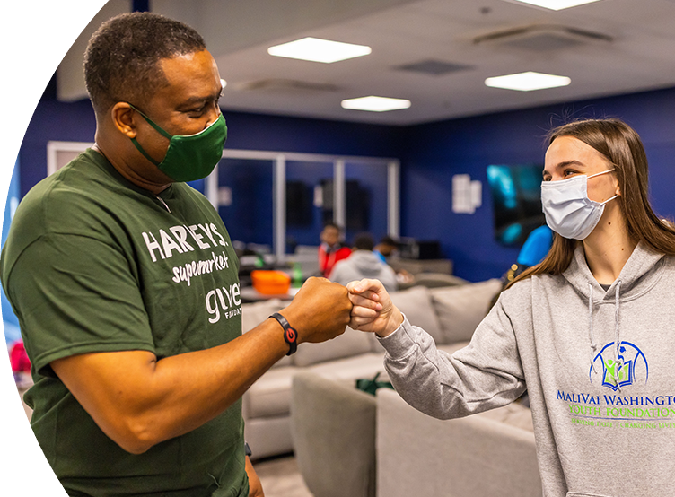 A young man in a Harveys Supermarket SEG Gives Foundation shirt and young woman in a MaliVai Washington grey sweatshirt wearing masks fist bumping.