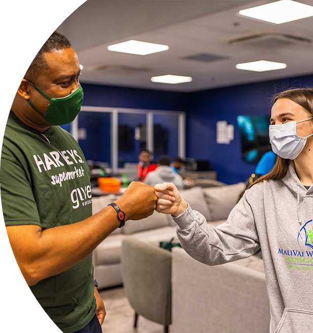 A young man in a Harveys Supermarket SEG Gives Foundation shirt and young woman in a MaliVai Washington grey sweatshirt wearing masks fist bumping.